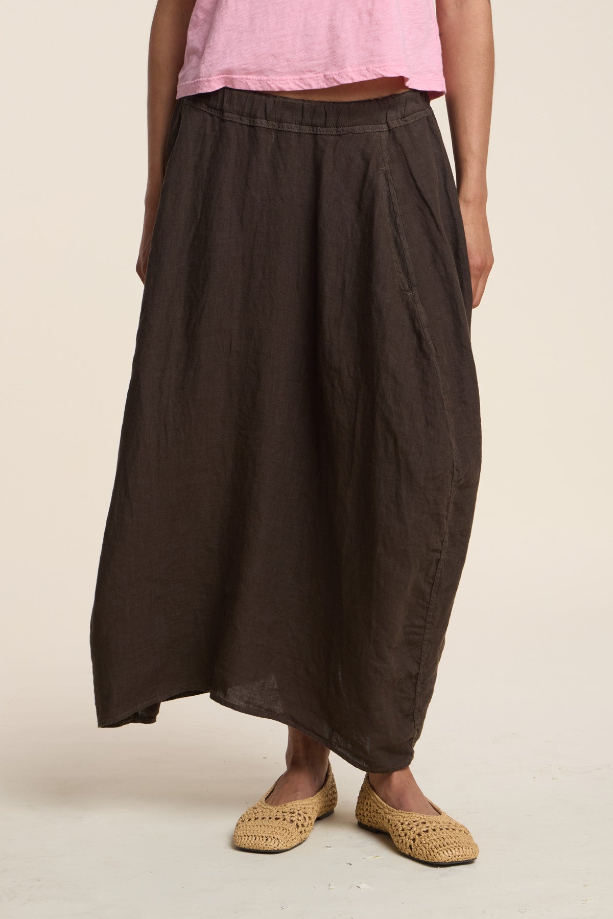 A person wears the FAE LINEN SKIRT by Velvet by Graham & Spencer—a loose, dark brown, A-line maxi linen skirt—with a pink top and woven beige slip-on shoes, standing against a plain background.-41460565606593