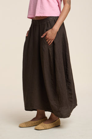 Someone wearing a pink short-sleeve top, the FAE LINEN SKIRT by Velvet by Graham & Spencer in dark brown with pockets and an A-line silhouette, and woven beige flats, stands against a plain background.