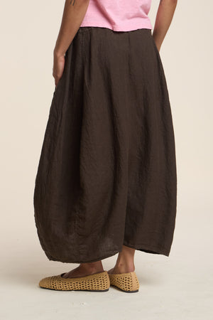 A person wearing a pink top, Velvet by Graham & Spencer's FAE LINEN SKIRT in wide brown A-line linen, and woven slip-on shoes stands against a plain background.
