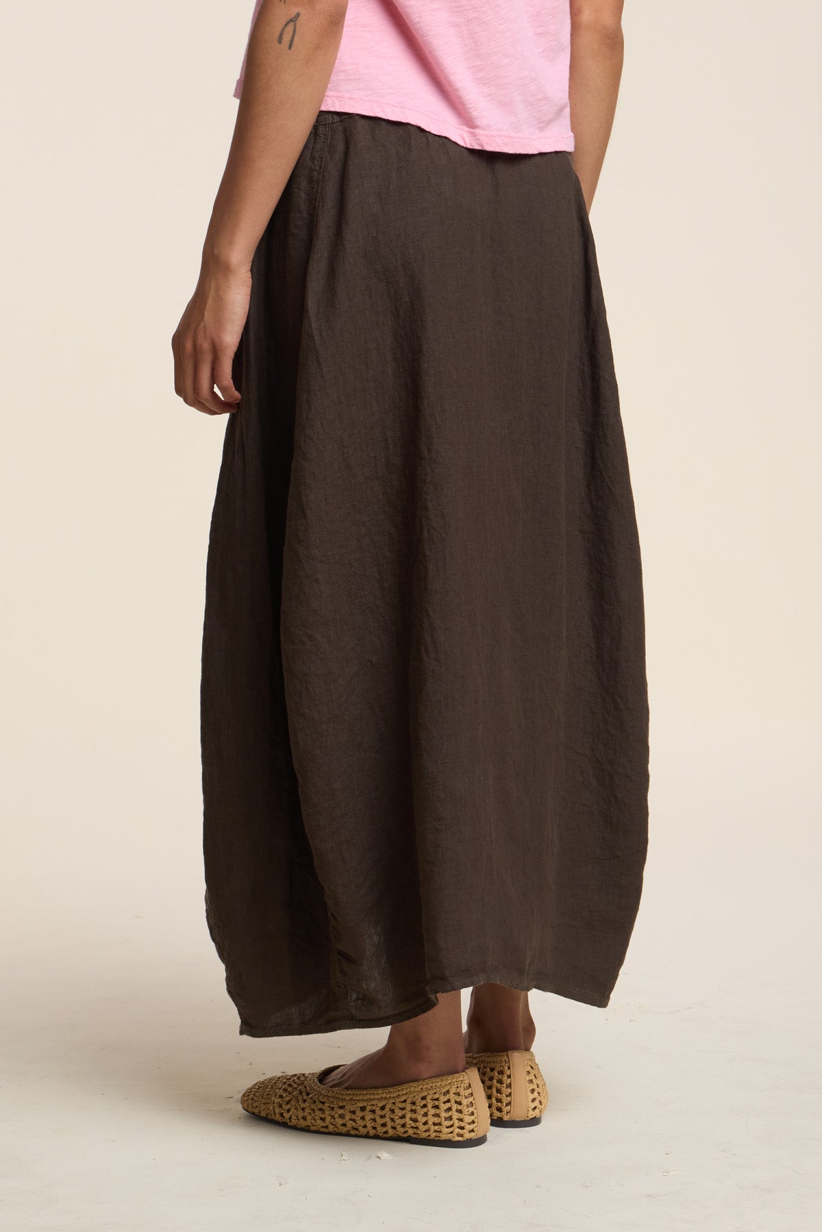 A person stands against a plain light background wearing a pink shirt, the FAE LINEN SKIRT by Velvet by Graham & Spencer in loose dark brown A-line linen, and woven beige shoes.-41460565737665