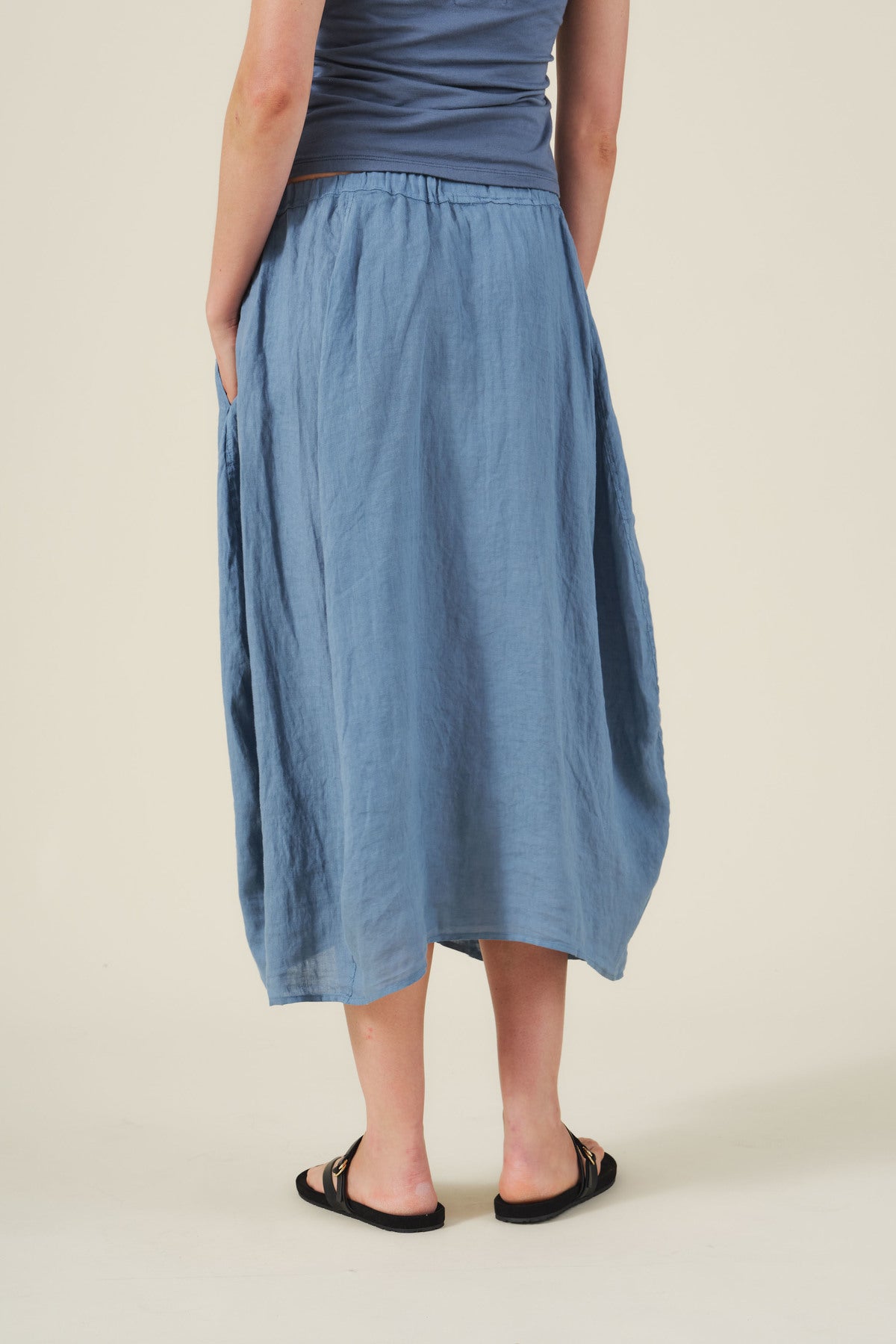 A person stands with their back to the camera, wearing the FAE LINEN SKIRT by Velvet by Graham & Spencer in blue, paired with a grey sleeveless top and black sandals.-41384932049089