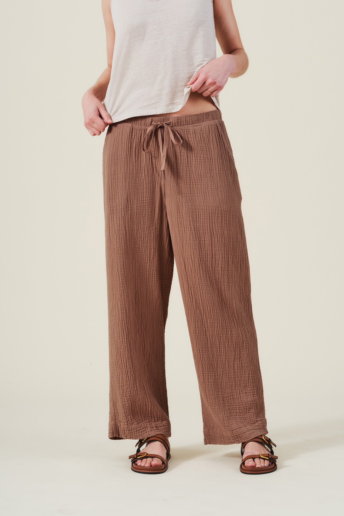 A person wears the FRANNY PANT by Velvet by Graham & Spencer—brown, wide-leg cotton gauze pants with a pull-on waistband—paired with a sleeveless beige top and brown sandals, standing against a plain light background.-41527344922817