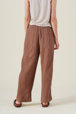 A person stands with back to camera wearing a sleeveless beige shirt, sandals, and the FRANNY PANT by Velvet by Graham & Spencer—loose, wide leg brown cotton gauze pants with a pull-on waistband.