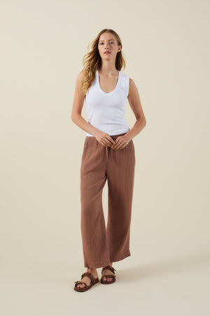 A woman stands against a plain background wearing a white sleeveless top, brown Velvet by Graham & Spencer FRANNY PANT with a comfortable pull-on waistband, and brown sandals.