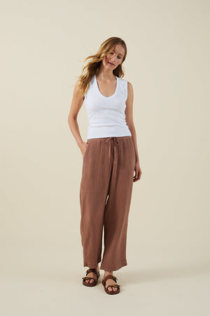 A woman stands against a plain background wearing a white sleeveless top, brown sandals, and the FRANNY PANT by Velvet by Graham & Spencer—wide leg cotton gauze pants with a pull-on waistband and one hand in her pocket.