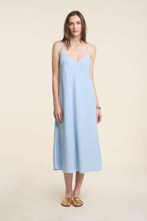 A woman wears the HOPE HEAVY LINEN DRESS by Velvet by Graham & Spencer, a light blue linen dress with adjustable straps, paired with yellow sandals against a beige background.