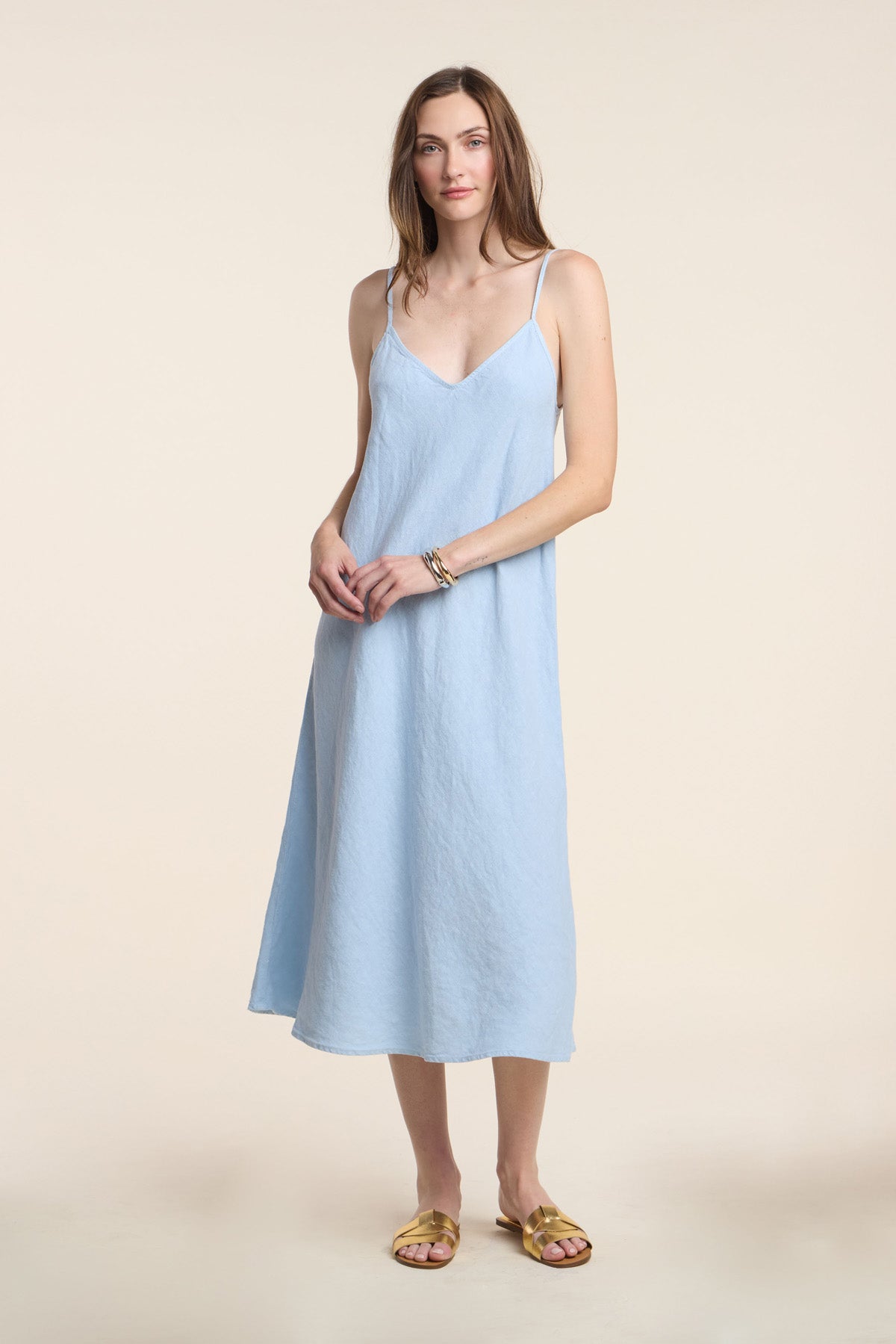 A woman stands against a plain beige background wearing the HOPE HEAVY LINEN DRESS by Velvet by Graham & Spencer in light blue with adjustable straps, paired with gold sandals.-41371937505473