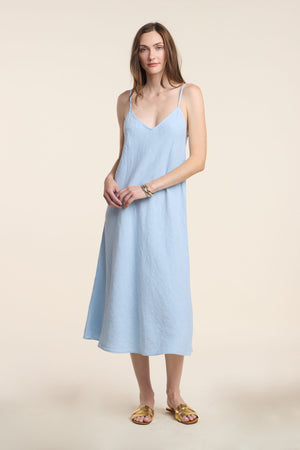 A woman stands against a plain beige background wearing the HOPE HEAVY LINEN DRESS by Velvet by Graham & Spencer in light blue with adjustable straps, paired with gold sandals.