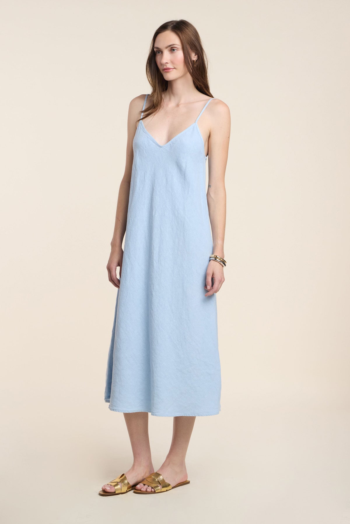 A woman with light brown hair stands against a beige background wearing the HOPE HEAVY LINEN DRESS by Velvet by Graham & Spencer—a sleeveless, light blue, ankle-length midi dress with adjustable straps—paired with gold sandals and bracelets.-41371937571009