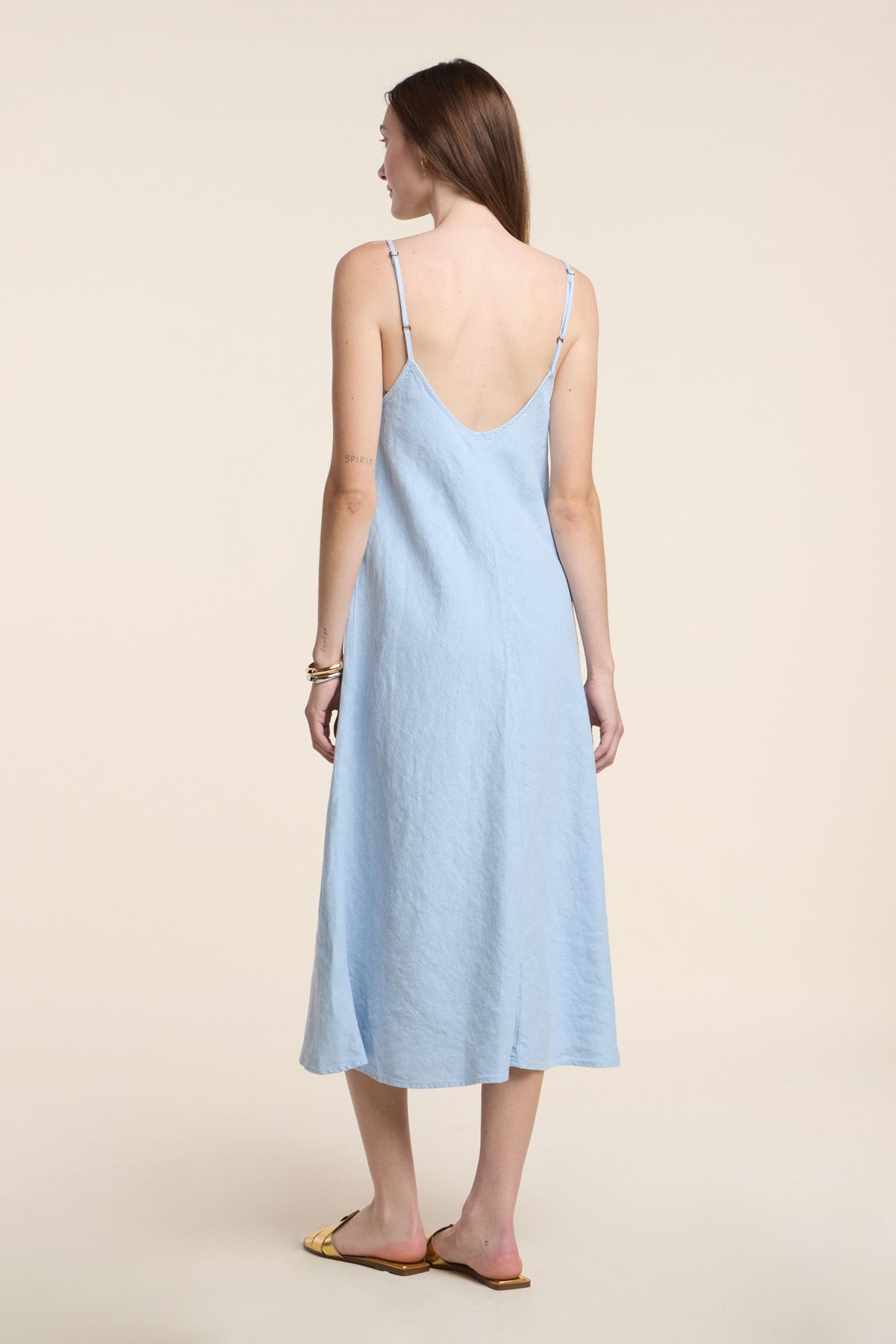 A woman faces away in the HOPE HEAVY LINEN DRESS by Velvet by Graham & Spencer, a light blue women’s midi dress with adjustable straps, paired with tan flat sandals against a plain light background.