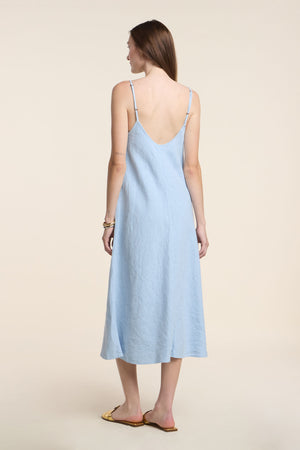 A woman faces away in the HOPE HEAVY LINEN DRESS by Velvet by Graham & Spencer, a light blue women’s midi dress with adjustable straps, paired with tan flat sandals against a plain light background.
