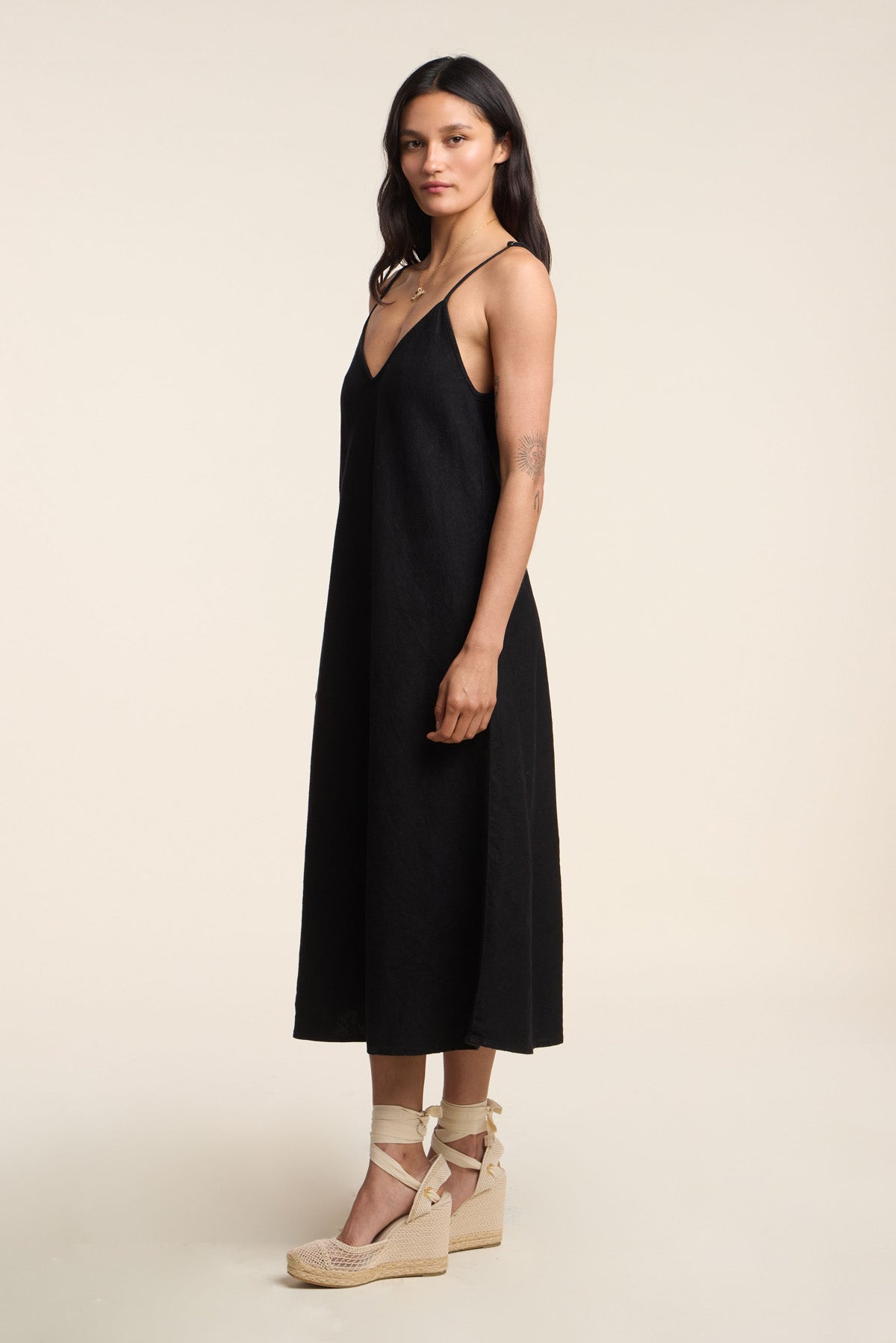 A woman stands in profile wearing the HOPE HEAVY LINEN DRESS by Velvet by Graham & Spencer, a sleeveless black midi dress, with beige wedge sandals against a plain light background.-41371938750657