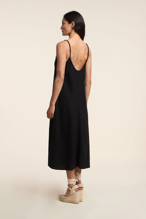 A woman stands sideways wearing the HOPE HEAVY LINEN DRESS by Velvet by Graham & Spencer—a sleeveless, black linen midi with a low back—paired with beige wedge sandals, creating a breathable summer-ready look against a plain background.