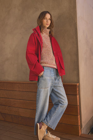 A woman stands indoors against a wooden bench and neutral wall, effortlessly styled for cold weather in the HUNTER PARKA by Velvet by Graham & Spencer, featuring cotton canvas construction with oversized front patch pockets, layered over a patterned sweater, wide-leg jeans, and beige sneakers.