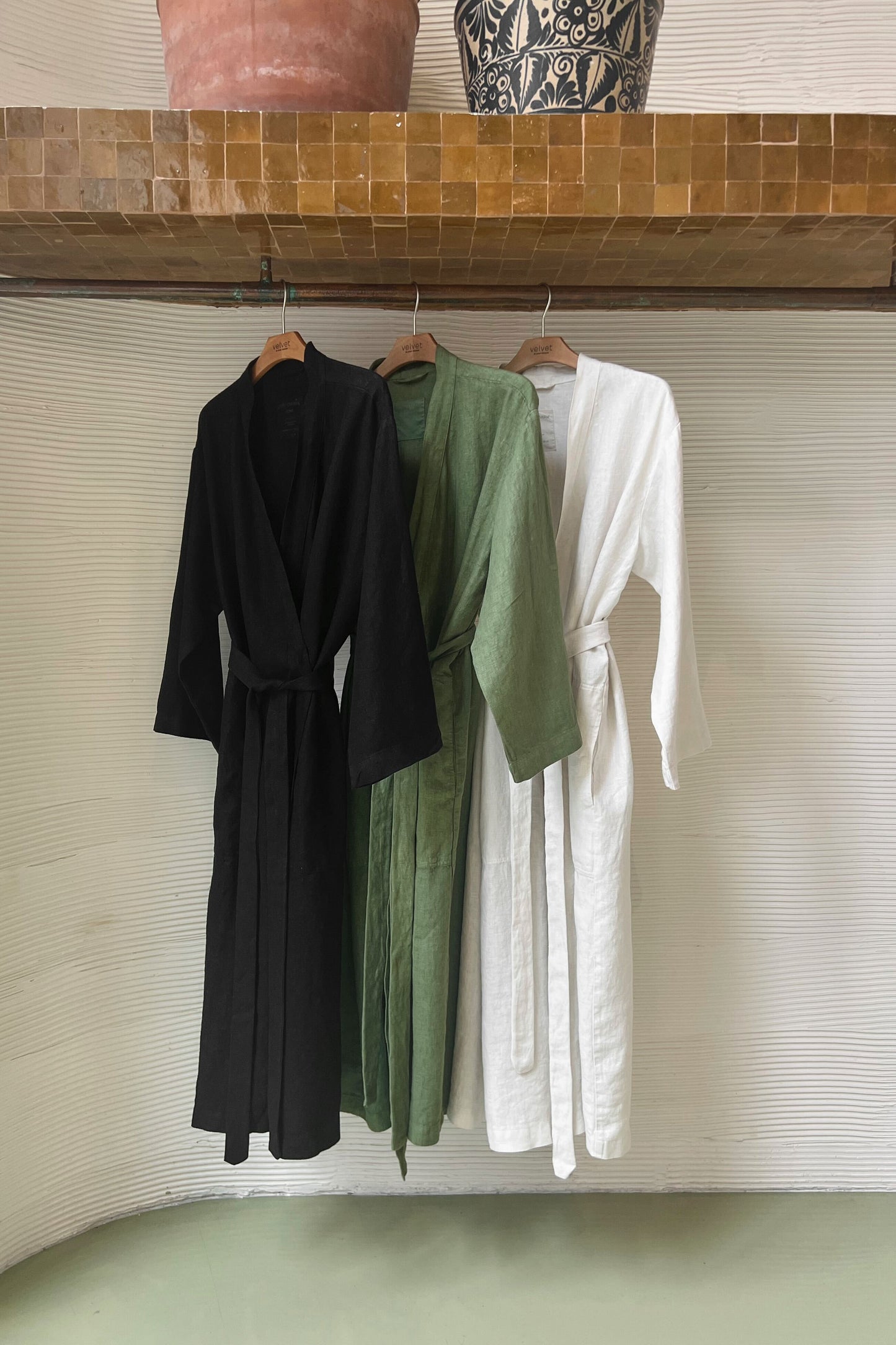 Jenny Graham Linen Robe in black on hanger near textured wall with robe in basil and beach-26312948318401