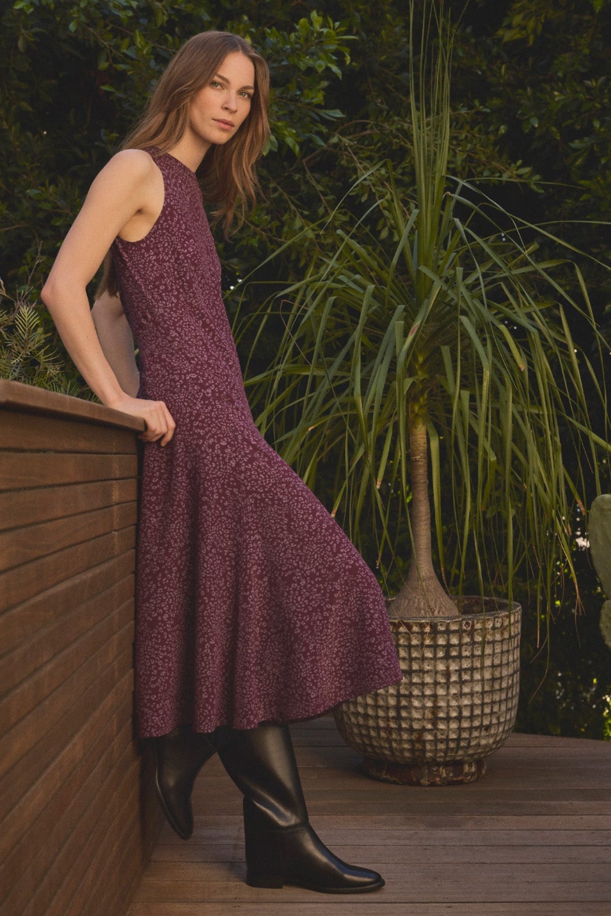 A woman wearing the INGRID SILK DRESS by Velvet by Graham & Spencer—a sleeveless purple silk dress with a flattering silhouette—pairs it with black boots as she stands on a wooden deck beside a large potted plant, green foliage in the background.-42092453757121
