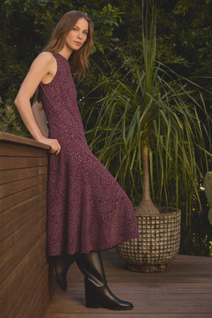 A woman wearing the INGRID SILK DRESS by Velvet by Graham & Spencer—a sleeveless purple silk dress with a flattering silhouette—pairs it with black boots as she stands on a wooden deck beside a large potted plant, green foliage in the background.