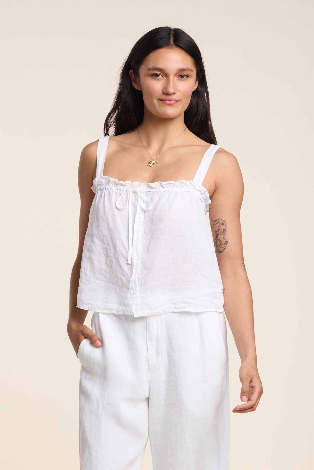 A woman faces forward against a plain background, wearing the IZZIE LINEN TANK TOP by Velvet by Graham & Spencer—a white woven linen tank with ruffle details—paired with white pants, one hand in her pocket.