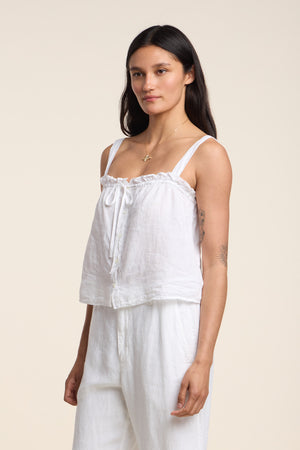 A woman stands slightly to the side in the IZZIE LINEN TANK TOP by Velvet by Graham & Spencer, featuring an adjustable fit and paired with matching white pants against a plain light background.