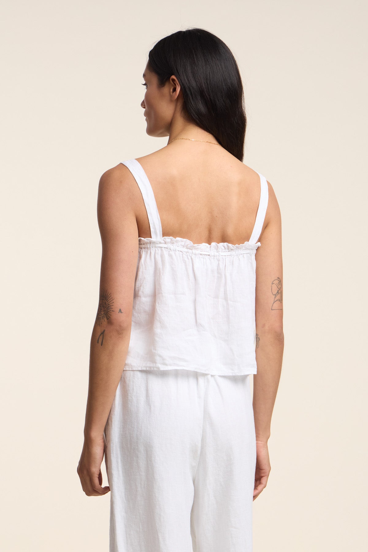 A woman with long dark hair faces away, wearing the IZZIE LINEN TANK TOP by Velvet by Graham & Spencer with white pants, set against a plain light-colored background.-41527046209729