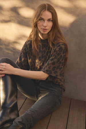 A woman with long brown hair sits on a wooden floor, wearing the stylish JANA TEE by Velvet by Graham & Spencer in a leopard print, paired with black jeans and knee-high black boots, as sunlight casts shadows on the wall behind her.