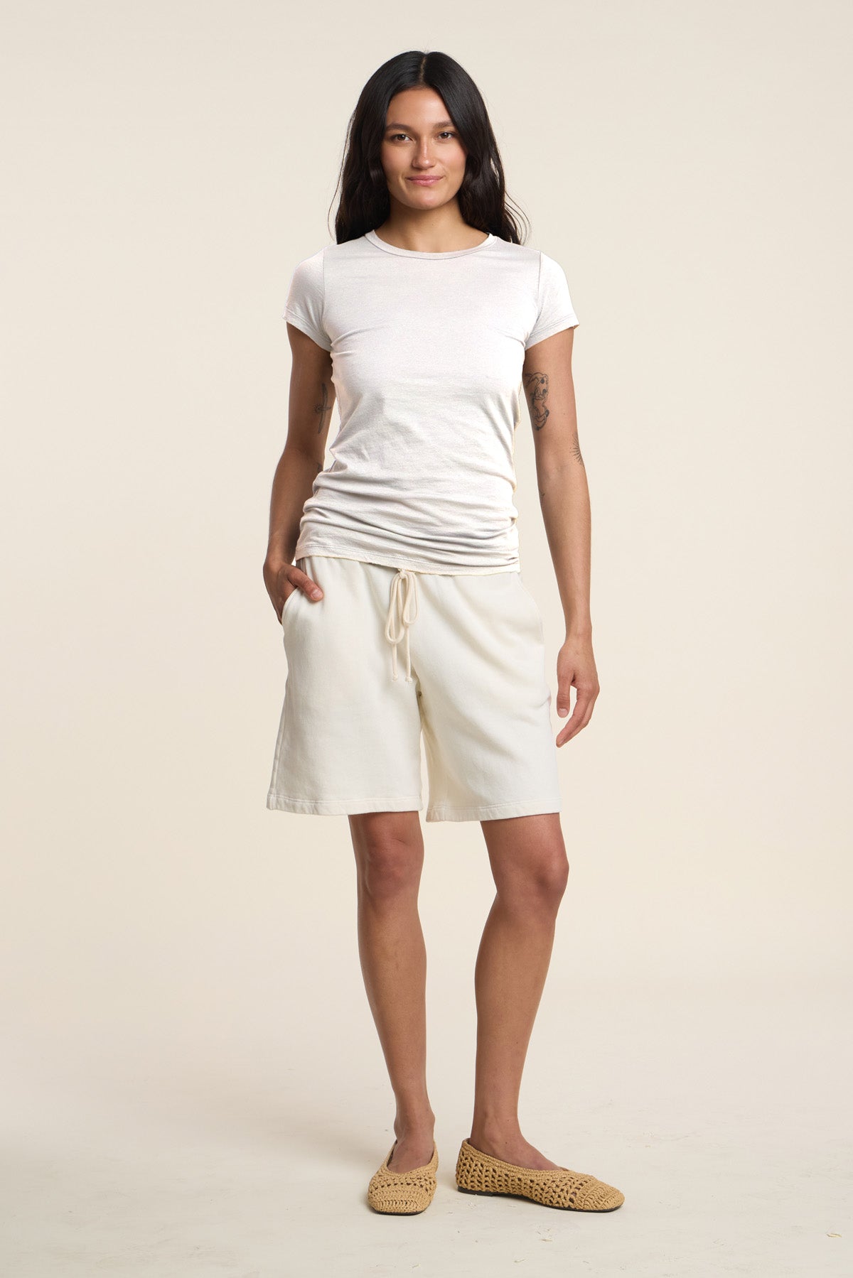 A woman stands facing forward against a plain light background, wearing the JEMMA TEE by Velvet by Graham & Spencer, cream drawstring shorts, and woven slip-on shoes.