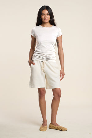 A woman stands facing forward against a plain light background, wearing the JEMMA TEE by Velvet by Graham & Spencer, cream drawstring shorts, and woven slip-on shoes.