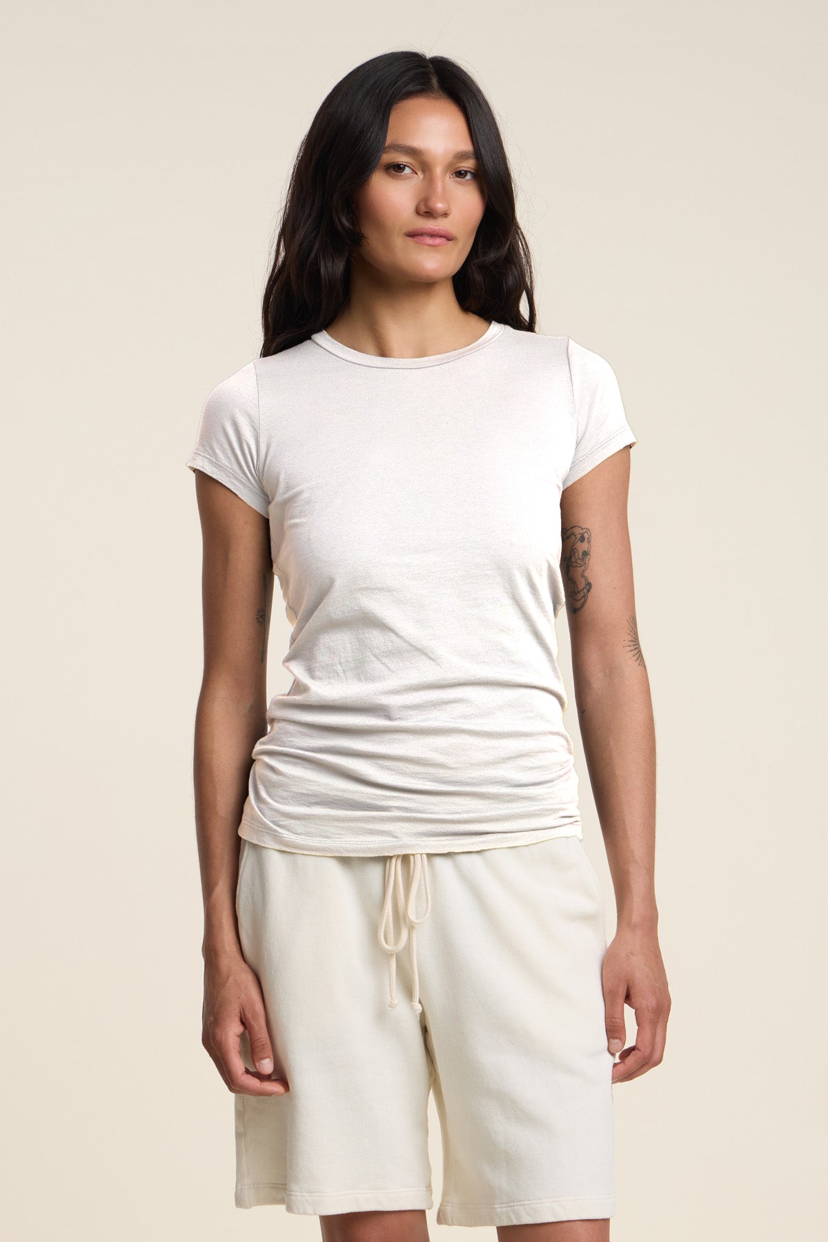 A person with long dark hair stands against a plain beige background, wearing the JEMMA TEE by Velvet by Graham & Spencer—an ultra-soft, fitted white crew neck tee—paired with white drawstring shorts.