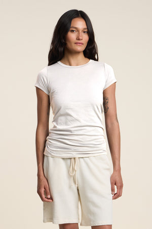 A person with long dark hair stands against a plain beige background, wearing the JEMMA TEE by Velvet by Graham & Spencer—an ultra-soft, fitted white crew neck tee—paired with white drawstring shorts.