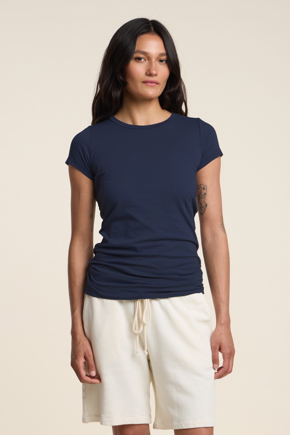 A woman with long dark hair wears the Velvet by Graham & Spencer JEMMA TEE in ultra-soft navy blue, paired with white drawstring shorts, standing against a plain light background.-41629735420097
