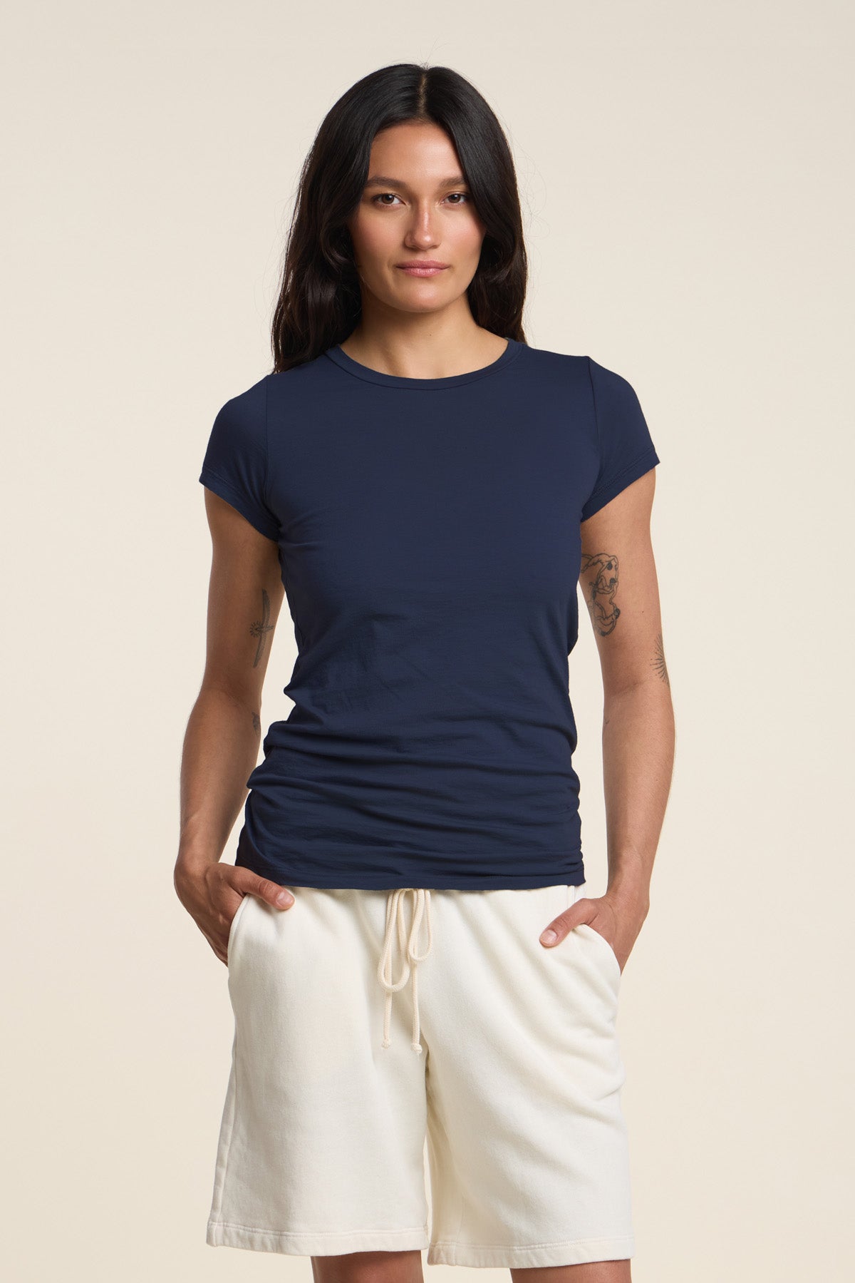 A person with long dark hair wears the Velvet by Graham & Spencer JEMMA TEE in navy and cream drawstring shorts, standing with hands in pockets against a plain background.