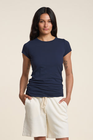 A person with long dark hair wears the Velvet by Graham & Spencer JEMMA TEE in navy and cream drawstring shorts, standing with hands in pockets against a plain background.