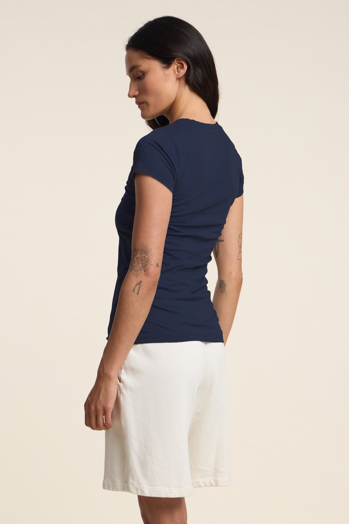 A woman with long dark hair and small arm tattoos stands sideways in a plain light-colored setting, wearing the JEMMA TEE by Velvet by Graham & Spencer in navy, paired with white shorts.
