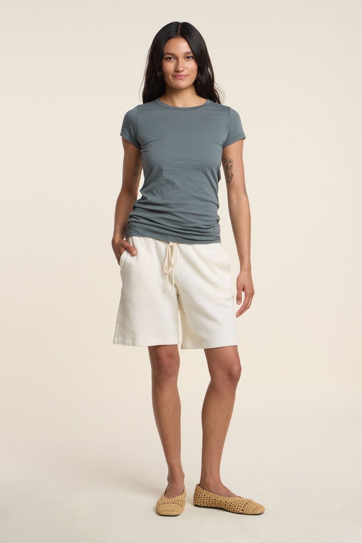 A person stands facing forward against a plain background, wearing the ultra-soft JEMMA TEE by Velvet by Graham & Spencer in gray, paired with white shorts and woven slip-on shoes, one hand in their pocket.-41371942846657