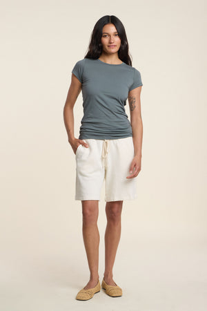 A woman faces forward against a light background, wearing a relaxed gray t-shirt, Velvet by Graham & Spencer LUE SWEATSHORT in off-white cotton fleece with a drawstring, and tan woven shoes.