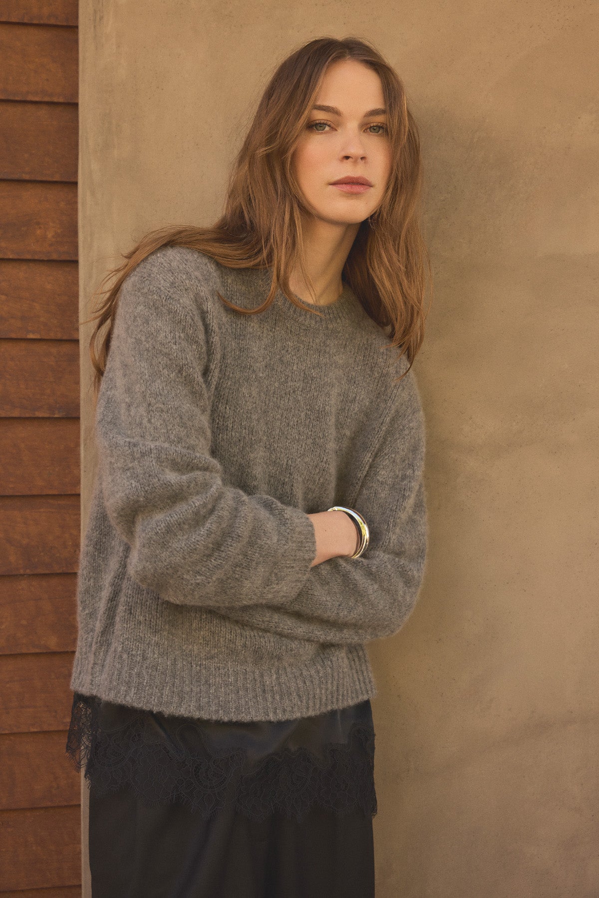 Wearing the JUNE SWEATER by Velvet by Graham & Spencer, a woman with long brown hair layers the cozy grey wool-cashmere piece over a black lace-trimmed top and black pants, standing against a textured beige wall with wooden siding to her left.