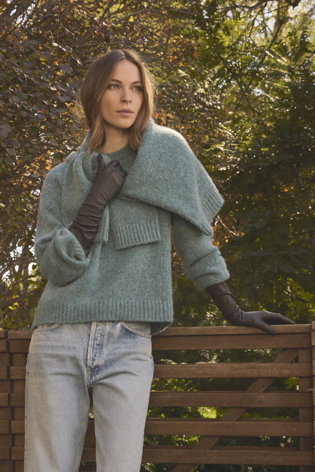 A woman stands outdoors by a wooden fence, surrounded by green foliage, wearing the JUNE SWEATER by Velvet by Graham & Spencer—a cozy, light blue wool-cashmere piece—paired with a matching scarf, jeans, and black gloves.-41997218939073