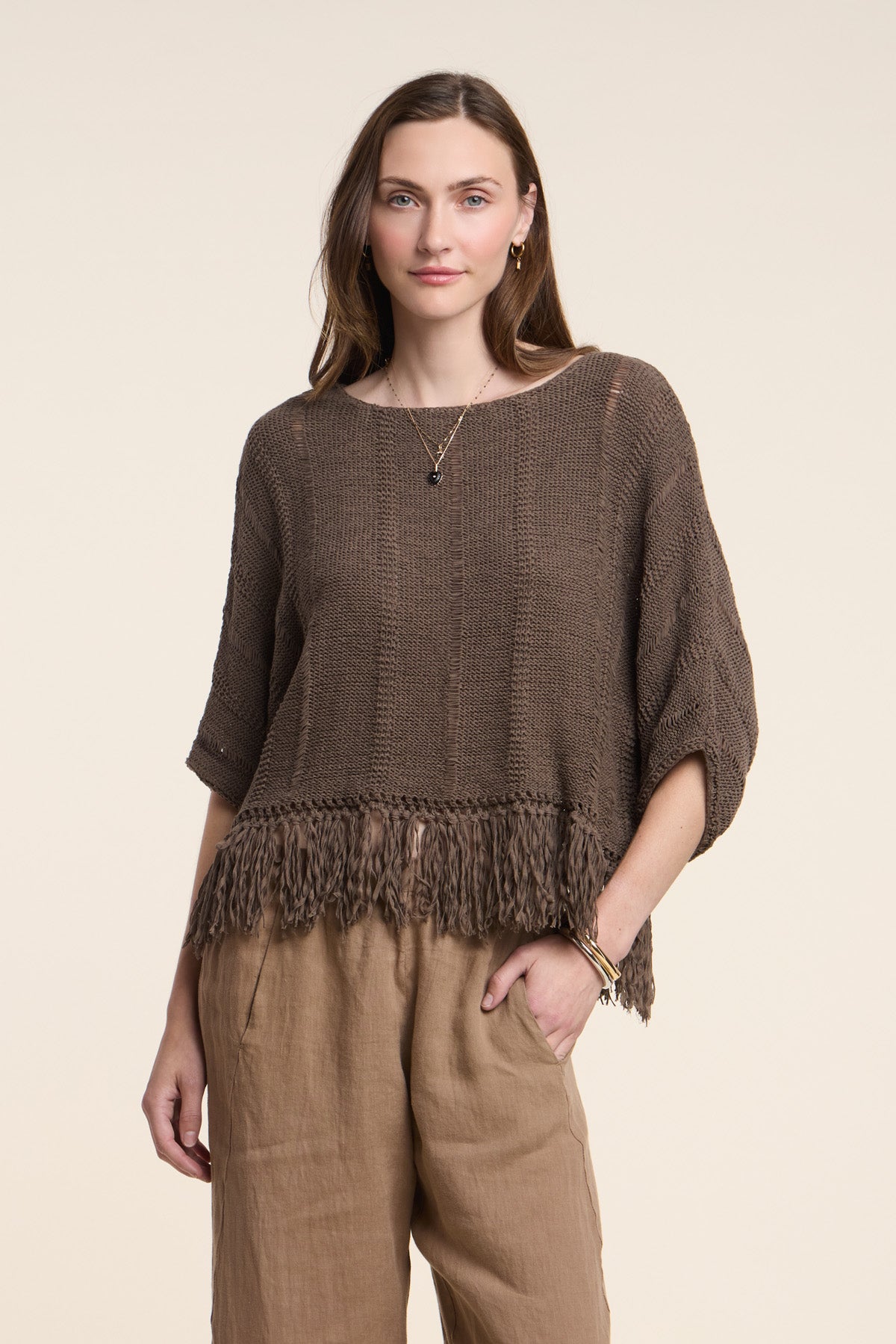 A woman stands facing forward with a hand in her pocket, wearing the KATARINA SWEATER by Velvet by Graham & Spencer—a loose brown cotton tape sweater with a fringe hem—paired with tan pants against a plain light background.