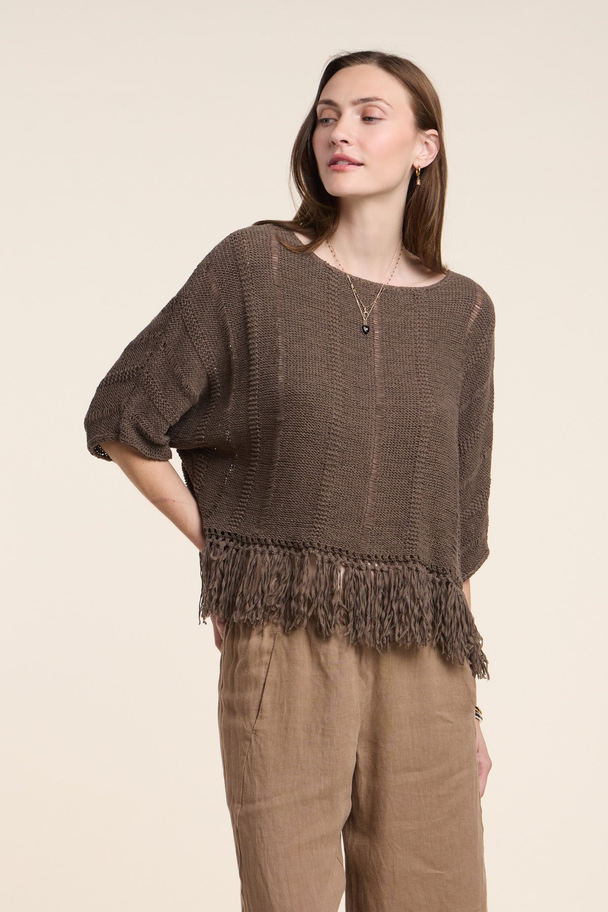 A woman wears the KATARINA SWEATER by Velvet by Graham & Spencer—an oversized cotton tape knit with a fringe hem—paired with light brown pants, standing against a plain background.-41527057875137