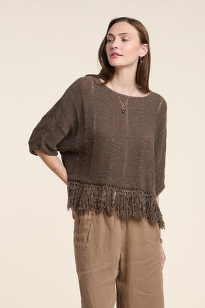 A woman wears the KATARINA SWEATER by Velvet by Graham & Spencer—an oversized cotton tape knit with a fringe hem—paired with light brown pants, standing against a plain background.