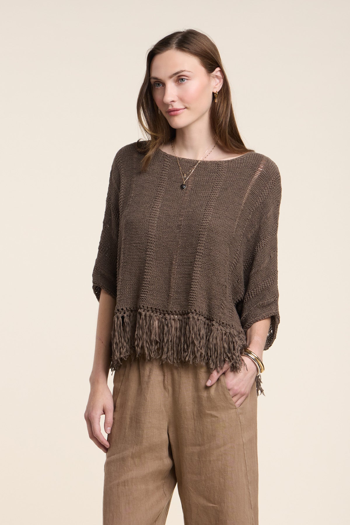 A woman wears the KATARINA SWEATER by Velvet by Graham & Spencer—a loose cotton tape sweater with a fringe hem—paired with light brown pants, standing against a plain background.