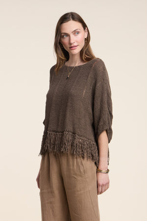 A woman wears the KATARINA SWEATER by Velvet by Graham & Spencer, a loose cotton tape sweater with a fringe hem, paired with wide beige pants against a plain light background.