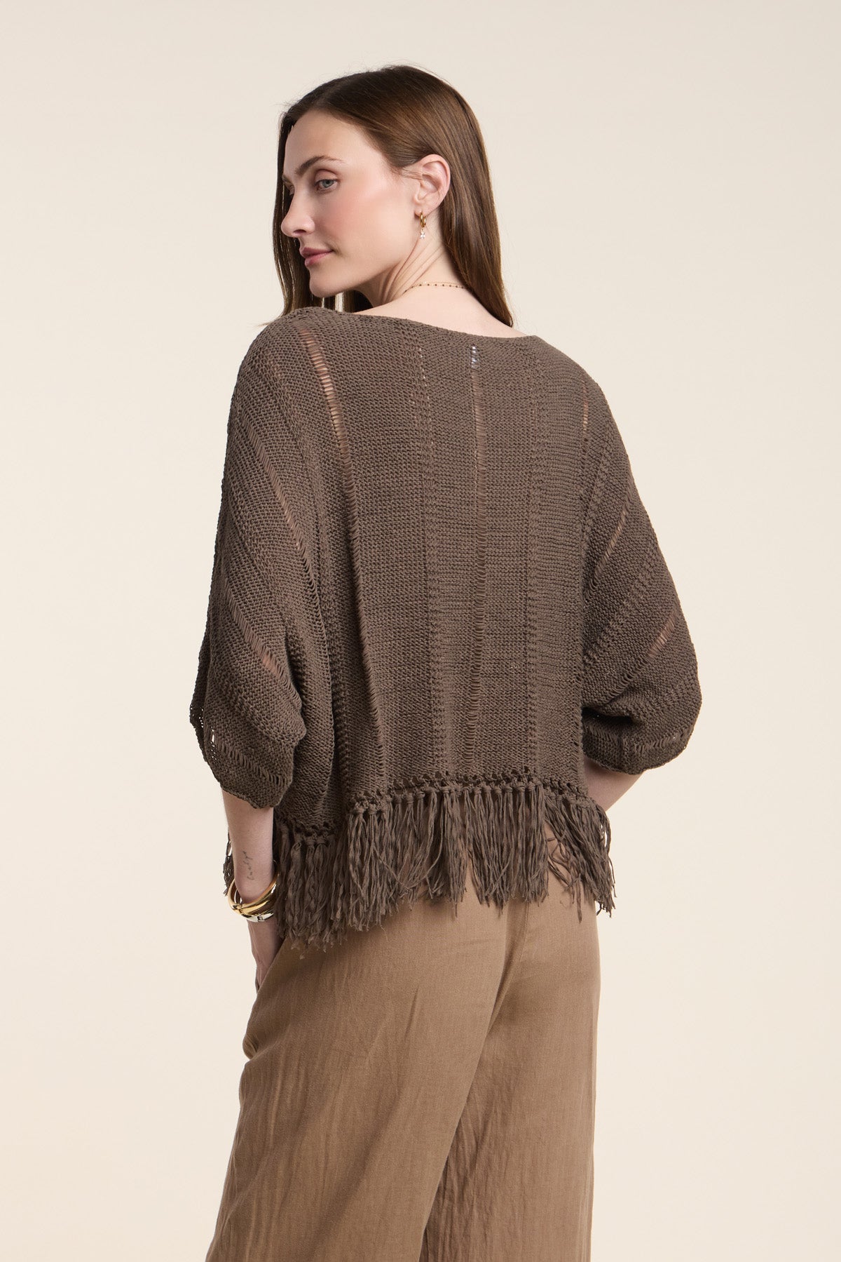 A woman stands with her back to the camera, wearing the oversized KATARINA SWEATER by Velvet by Graham & Spencer, featuring a brown knit design with a fringe hem and paired with wide-leg brown pants.