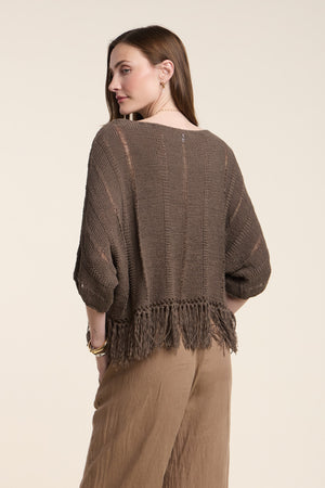 A woman stands with her back to the camera, wearing the oversized KATARINA SWEATER by Velvet by Graham & Spencer, featuring a brown knit design with a fringe hem and paired with wide-leg brown pants.