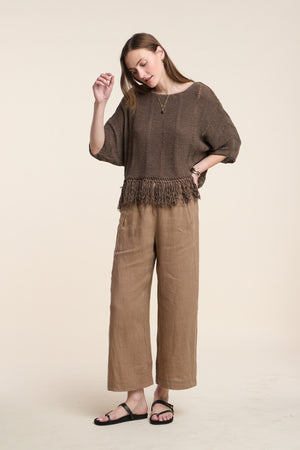 A woman stands against a plain beige background wearing relaxed straight leg LOLA LINEN PANT by Velvet by Graham & Spencer, paired with a loose brown knit top with fringe and black sandals.