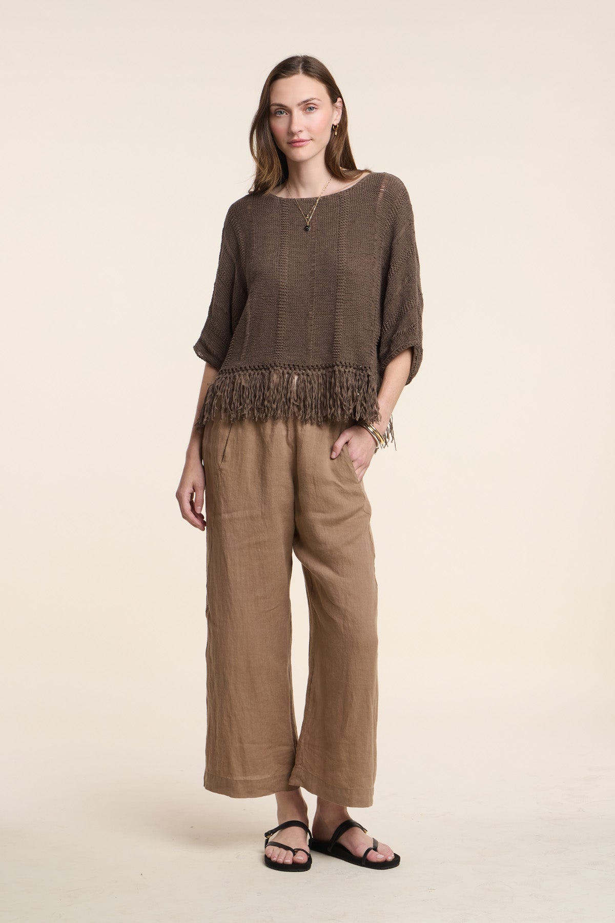 A woman stands against a plain light background in neutral tones, wearing a loose brown top with fringe hem, black sandals, simple jewelry, and the LOLA LINEN PANT by Velvet by Graham & Spencer.-41527057776833