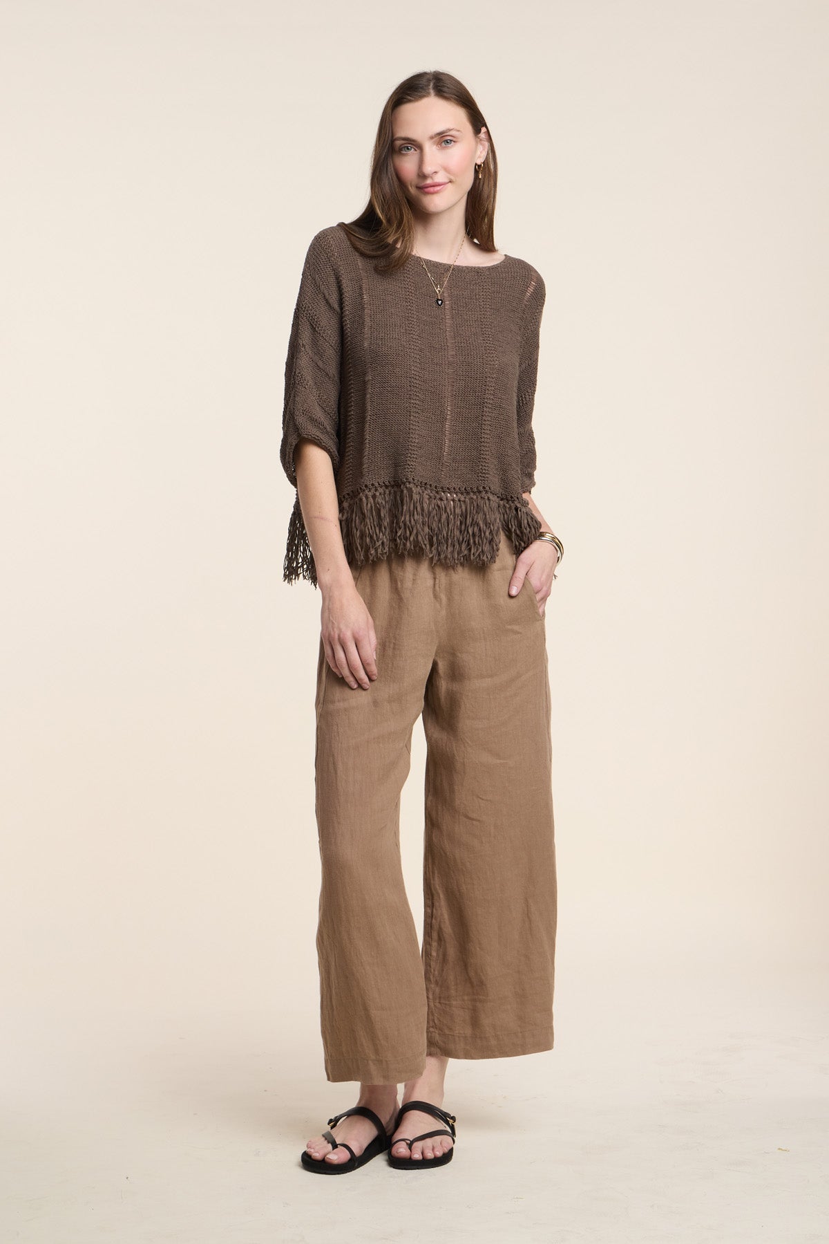 A woman stands against a plain background, wearing Velvet by Graham & Spencer’s LOLA LINEN PANT—a pair of loose linen pants with an elastic waistband—paired with a brown fringed sweater, black sandals, and a gold necklace. Her hair is down and one hand rests by her side.-41527057809601