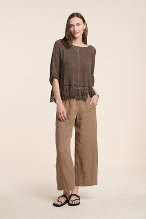 A woman stands against a plain background, wearing Velvet by Graham & Spencer’s LOLA LINEN PANT—a pair of loose linen pants with an elastic waistband—paired with a brown fringed sweater, black sandals, and a gold necklace. Her hair is down and one hand rests by her side.