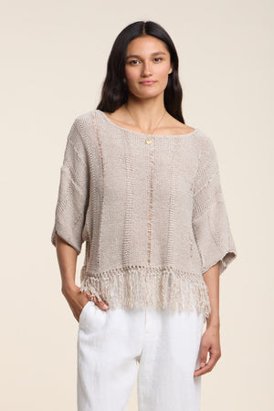 A woman in the KATARINA SWEATER by Velvet by Graham & Spencer—a loose-knit cotton tape sweater with a fringe hem—pairs it with white pants and stands with one hand in her pocket against a plain background.