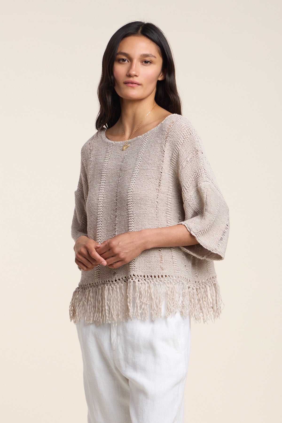 A woman stands against a plain off-white background wearing the KATARINA SWEATER by Velvet by Graham & Spencer—an oversized beige cotton tape sweater with a fringe hem—paired with white pants.